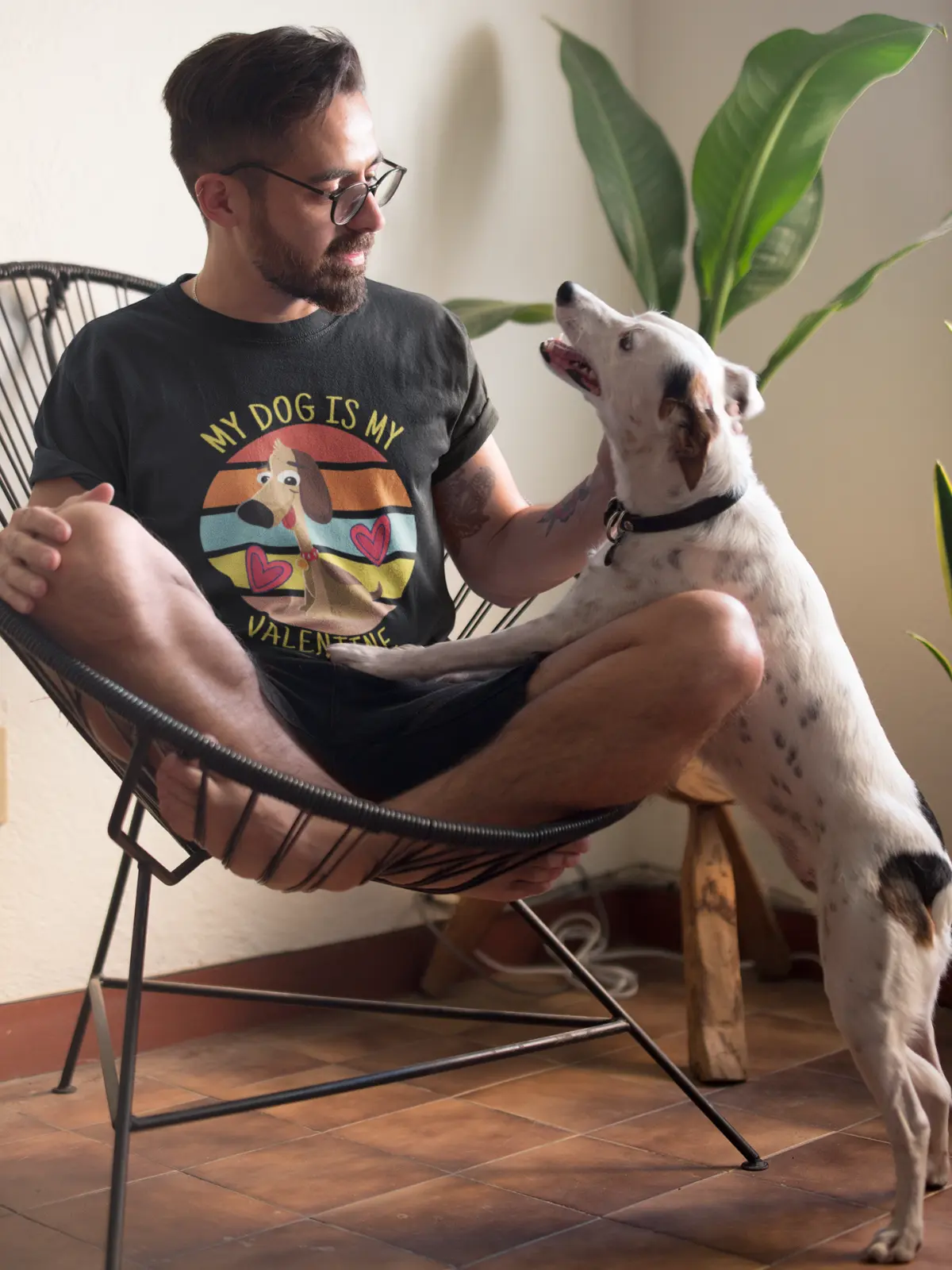 Mann mit Bart sitzt barfuß im Designerstuhl und trägt ein T-Shirt mit dem Aufdruck ‚My Dog is my Valentine‘, während er liebevoll mit seinem weißen Hund mit schwarzen Flecken interagiert – perfekte Szene für Hundeliebhaber.