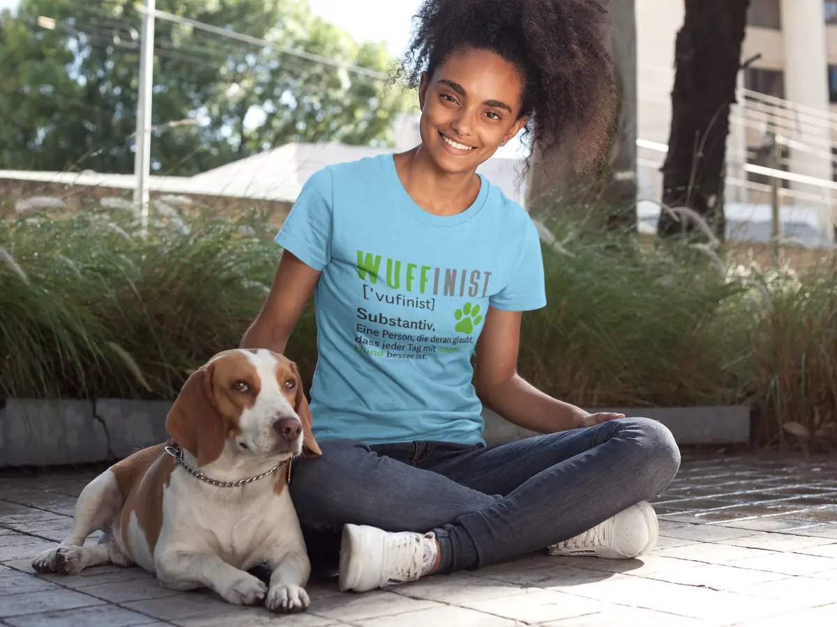 Lachende Frau im blauen T-Shirt mit Aufdruck ‚Wuffinist – Substantiv: Eine Person, die daran glaubt, dass jeder Tag besser ist mit einem Hund‘, sitzt mit Beagle auf einem gepflasterten Weg im Grünen.